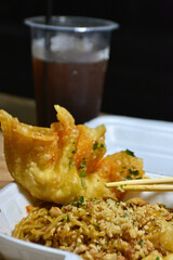 Close-up of fried wonton and seasoned noodles in a takeout container with a cold drink in the...