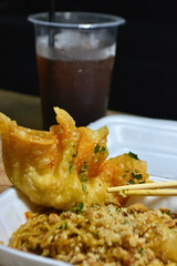 Close-up of fried wonton and seasoned noodles in a takeout container with a cold drink in the...