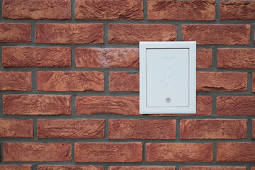 Exterior brick wall with an electrical service box that is white and has a grounding symbol and...