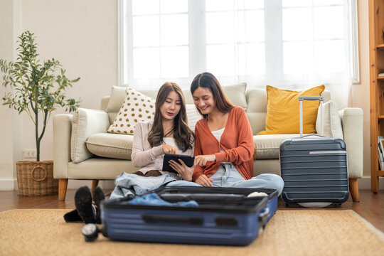 Two asian friends woman packing suitcase and choosing clothes together using tablet, planning a trip, checking fashion reviews or packing lists, travel preparation, content creator and travel blogger - Powered by Adobe