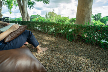 Barefoot woman enjoying digital detox by reading book outdoors at riverside hotel resort with...