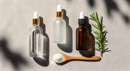 Flatlay featuring amber and clear glass skincare bottles with dropper tops, a wooden spoon filled with clay powder, and a sprig of rosemary. Placed on linen fabric with soft natural shadows.