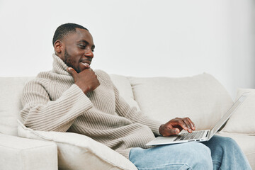 Young African man in cozy sweater enjoying online browsing on a laptop in a modern living room,...