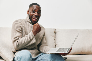 Happy African American man in a cozy sweater celebrating success with a laptop at home, conveying...