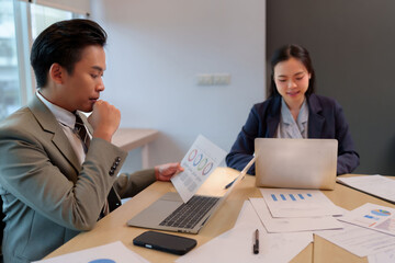 Obraz premium Asian adult male business professional reviews financial data charts reports using laptop with female colleague at office meeting desk