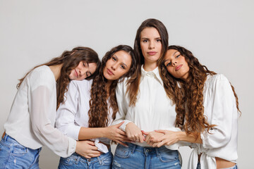 Four women with long wavy hair posing together wearing white blouses and denim jeans, showcasing...