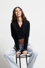 Confident young woman sitting on a stool with a bright smile, wearing a casual blazer with jeans in...