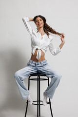 Cheerful woman in a white shirt sitting on a high stool, posing confidently in a studio setting