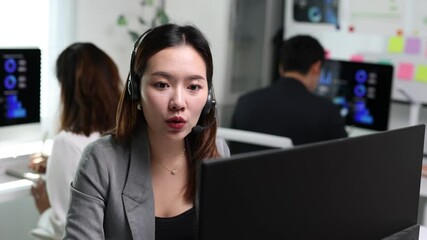 Professional Asian businesswoman working at computer, wearing headset while communicating with colleagues in modern office, collaborative workspace. - Powered by Adobe