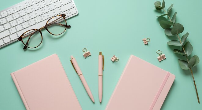Flat lay of office supplies including keyboard, notebooks, pens, and glasses on a mint green background.
