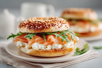 Close up of a bagel sandwich filled with cream cheese, smoked salmon, and fresh arugula on a white plate, set against a minimalistic backdrop