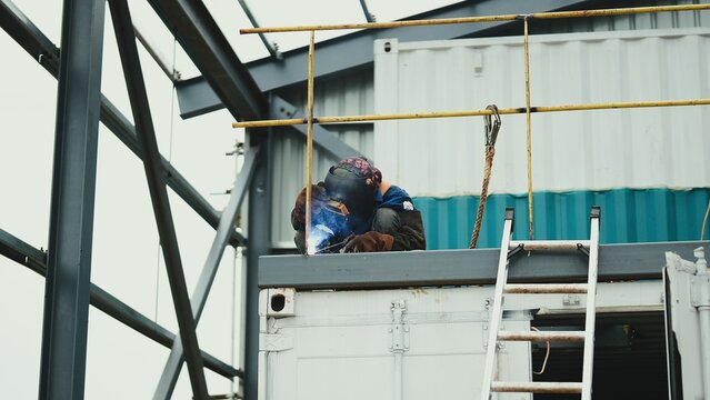 A construction worker performs welding on a steel structure at a high elevation, wearing proper protective gear including a welding helmet, gloves, and safety harness
