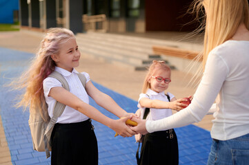 happy schoolgirls with backpacks receive apples from woman outdoors by school entrance. back-to-school theme with children in uniforms shows caring gesture and healthy lifestyle.