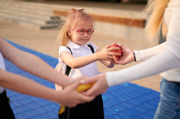 child receives apple from adult. young girl in red glasses at school receives fruit from teacher in...