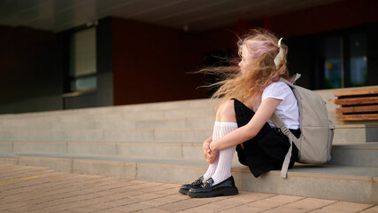 young girl with backpack sitting on outdoor school steps, reflecting in solitude. wind blowing...