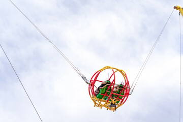 A catapult ride at an amusement park where two young women sit in a spherical cage against the sky, their long dark hair blowing in the wind. A thrilling ride for adults and teenagers.
