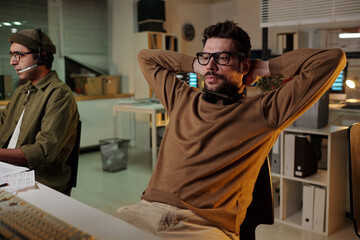 Caucasian man with headset leaning back in office chair relaxing while another man with headset...