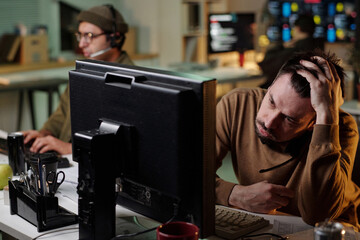 Caucasian man sitting at desk holding head in hand while using computer, with young adult man in...