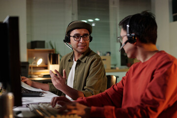 Young Asian man and Indian man wearing headsets sitting at desks operating computers, engaging in conversation while working in scam call center