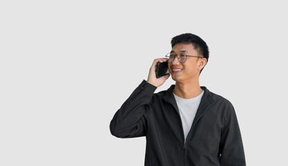 Young Asian man in his 20s wear glasses and wearing a black coat is talking on the phone standing on a gray background. Long-distance communication concept. Happy conversation. Smile on the face