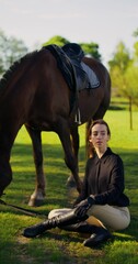 A stylish woman poses looking at the camera while sitting outdoors. The horse grazes standing next...