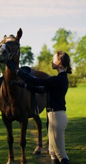 A stylish woman poses looking at the camera standing next to a horse and holding the reins
