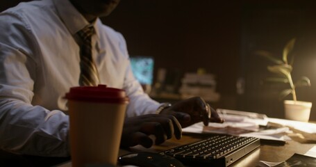 A businessman uses a computer while sitting at his desk in a dark office. Close-up of his hands,...