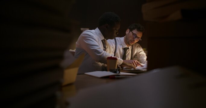 The two detectives work together. They use a computer and study documents while sitting at their desk in the office.