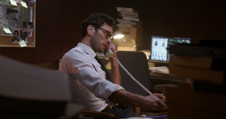 The detective is talking on the phone with a serious look. He's sitting at his desk in the office.