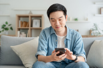 Smiling handsome young Asian man sitting on cozy couch, using funny mobile apps in living room. man...