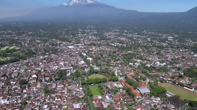 A cityscape of Salatiga with urban buildings in the foreground, framed by the majestic Mount Merbabu rising peacefully in the background under a clear sky.