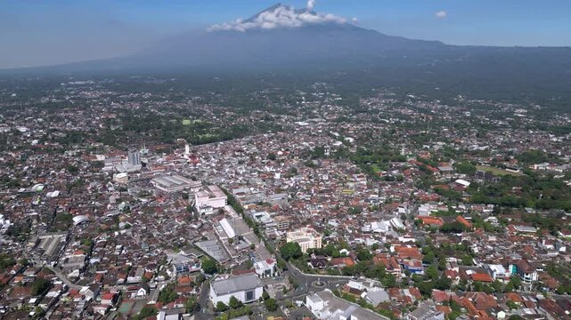 A cityscape of Salatiga with urban buildings in the foreground, framed by the majestic Mount Merbabu rising peacefully in the background under a clear sky.