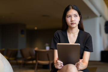 Fototapeta premium Young Asian woman sitting in coffee shop wearing black dress using tablet computer