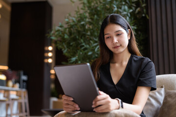 Obraz premium Young Asian woman sitting in coffee shop wearing black dress using tablet computer