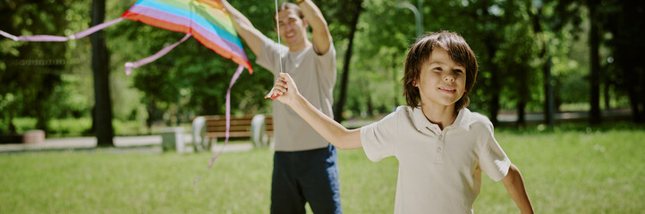 Biracial boy running with colorful kite while middle aged Dad standing behind holding kite string...