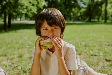 Portrait of biracial boy child sitting outdoors in park holding sandwich with both hands, eating lunch looking at camera surrounded by green grass and trees in background