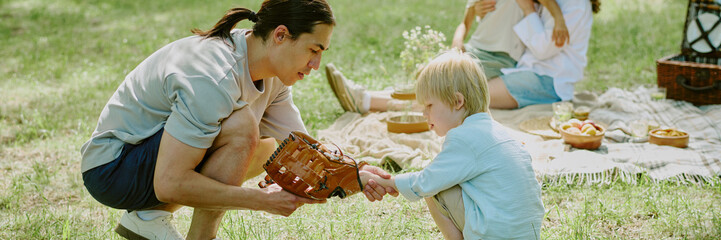 Biracial child learning to play baseball with young adult Asian father in park, while his mother...