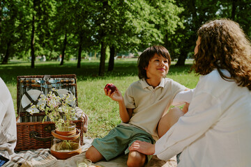 Woman sitting on picnic blanket interacting with biracial boy holding apple, picnic basket with...
