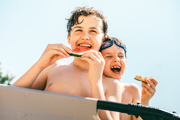 Two happy boys laugh and enjoy juicy watermelon slices on a sunny summer day at the swimming pool