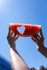 A hand holds a piece of watermelon with a heart-shaped cutout against a bright blue sky. The vivid...