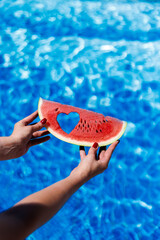 A hand holds a piece of watermelon with a heart-shaped cutout against a swimming pool. The vivid...