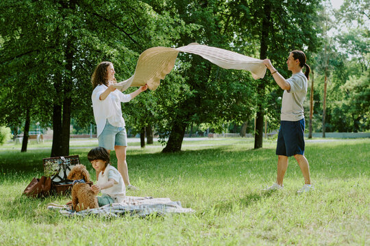 Caucasian middle aged woman and Asian young adult man spreading blanket on grass, while biracial child sitting on picnic blanket petting brown dog in green park setting - Powered by Adobe