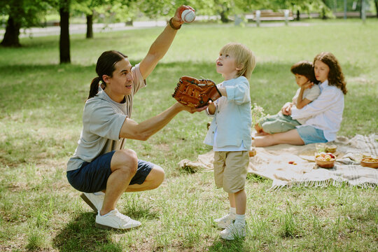 Asian young adult man teaching child boy to catch baseball in park, while Caucasian middle aged woman and child boy sitting on picnic blanket in background - Powered by Adobe