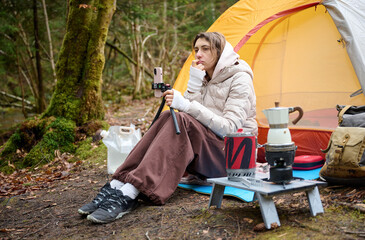 A beautiful girl blogger sits near a tent in the forest and films herself on a smartphone camera....