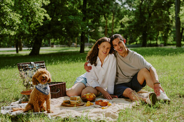 Portrait of young multiethnic couple sitting on picnic blanket in park, smiling at camera with...