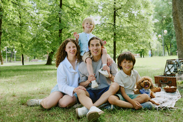 Fototapeta premium Diverse parents with two children and brown dog sitting on grass in park, smiling and relaxing together, young boy sitting on man's shoulders, picnic basket nearby