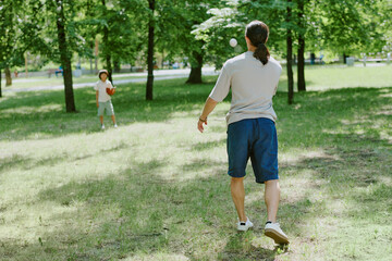 Father playing catch with young child in green park, man throwing baseball toward child holding...