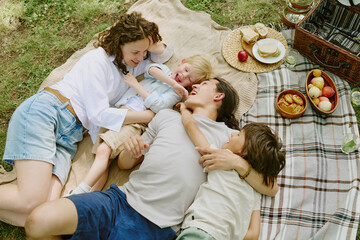 Caucasian young woman, Asian young man, two biracial boys lying on picnic blanket in park laughing and hugging near picnic basket and food