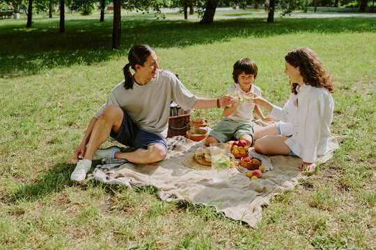 Multiethnic family with biracial child sitting on picnic blanket in park sharing food and drinks, smiling and interacting outdoors on sunny day