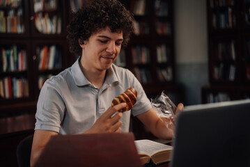 Student eating pastry while studying in library with laptop and book